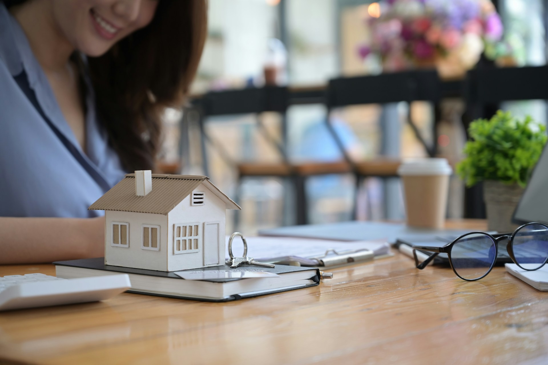 A model house, keys, and mortgage paperwork are on a wooden table; a woman sits in the background, partially visible and smiling at the high commitment of her new home loan.